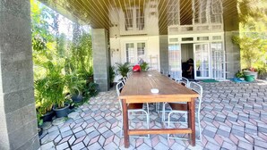 Un espacio comedor al aire libre con techo, una larga mesa de madera y sillas de metal, rodeado de plantas en macetas y un suelo de baldosas.