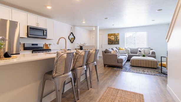 A modern kitchen with white cabinets, a microwave, and a stove. There is a dining area with a bar stool and a sofa. The room has a large window and a painting on the wall.