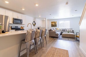 Una cocina moderna con armarios blancos, un microondas y una estufa. Hay un comedor con un taburete de bar y un sofá. La habitación tiene un ventanal grande y un cuadro en la pared.