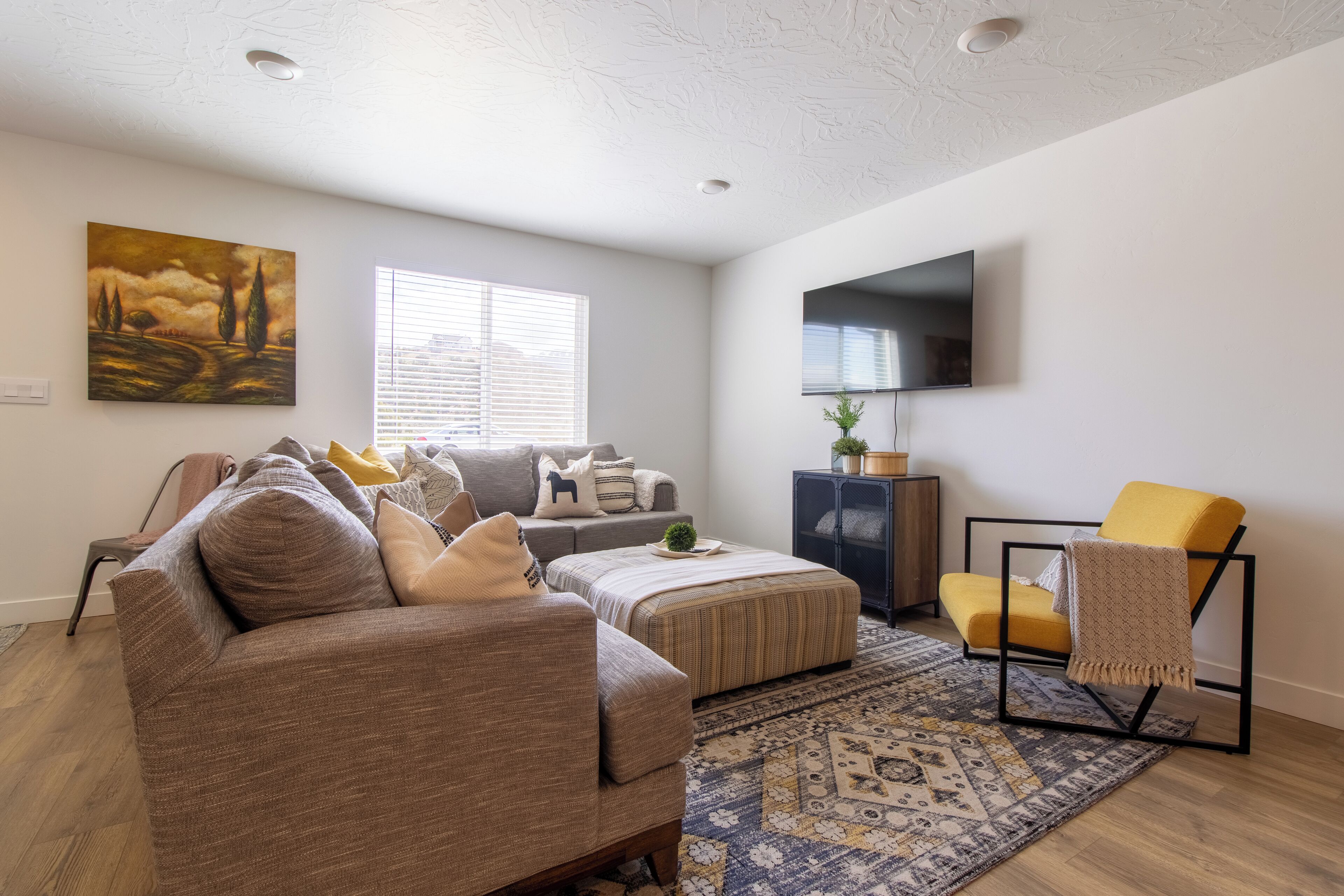 A modern living room with a sofa, ottoman, and armchair, a flat-screen TV mounted on the wall, and a patterned rug.