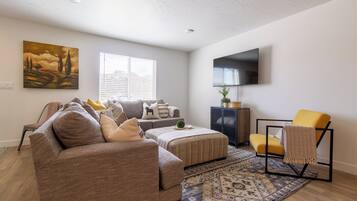 A modern living room with a sofa, ottoman, and armchair, a flat-screen TV mounted on the wall, and a patterned rug.