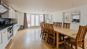 Una cocina moderna con armarios blancos, una mesa de comedor de madera y sillas. Hay un ventanal con cortinas y una vista de un edificio de ladrillo.