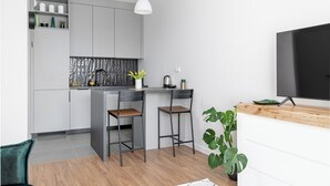 A modern kitchen with grey cabinets, a black countertop, two bar stools, a hanging pendant light, and a TV on a wooden shelf. - Apartament Ludwiki (Warsaw)