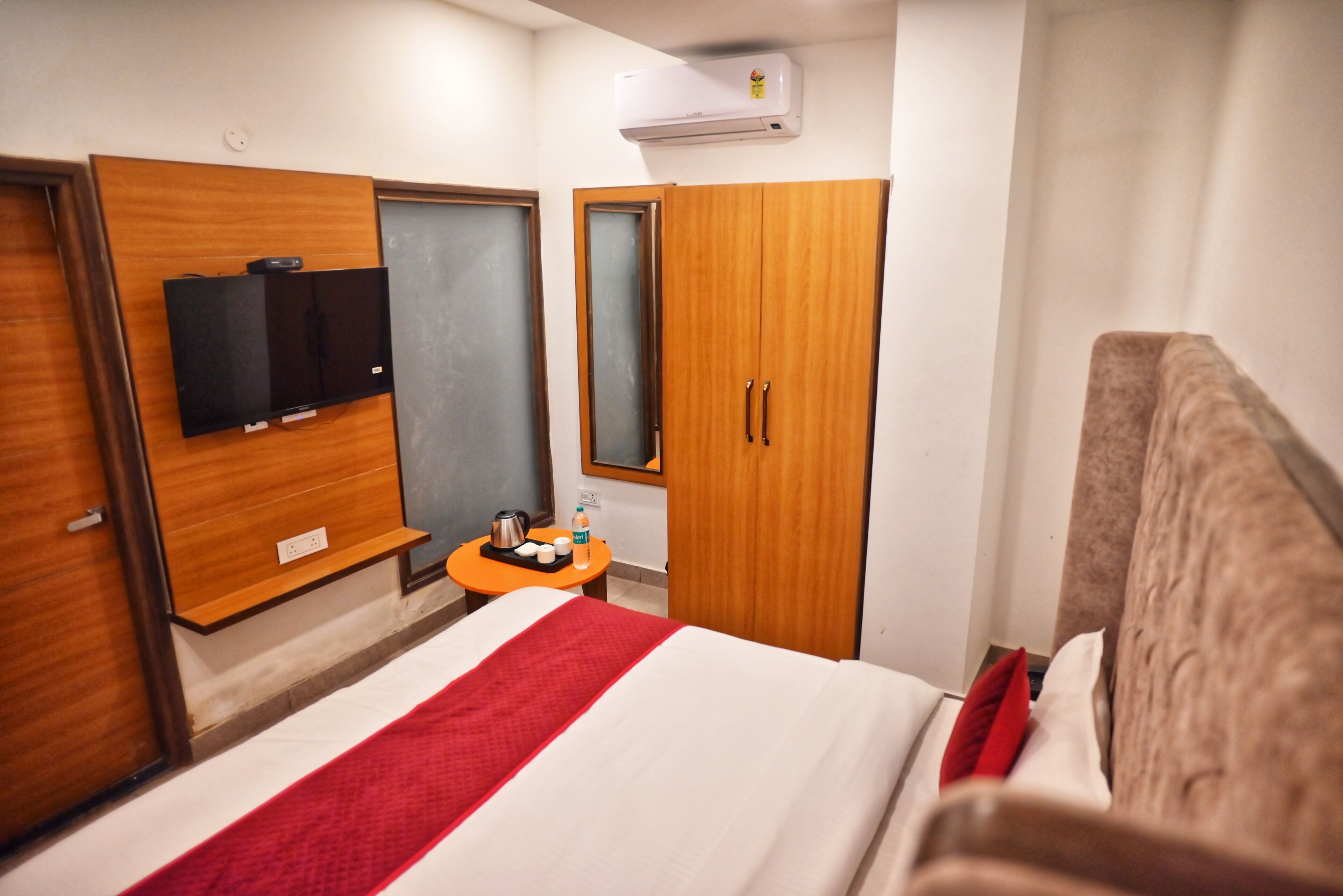 A hotel room with a bed, a wooden wardrobe, a small table with a kettle and cups, and a flat-screen TV mounted on the wall.