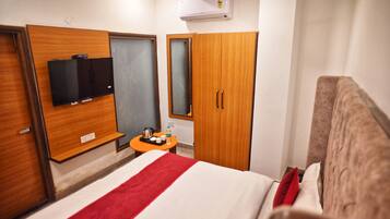 A hotel room with a bed, a wooden wardrobe, a small table with a kettle and cups, and a flat-screen TV mounted on the wall.