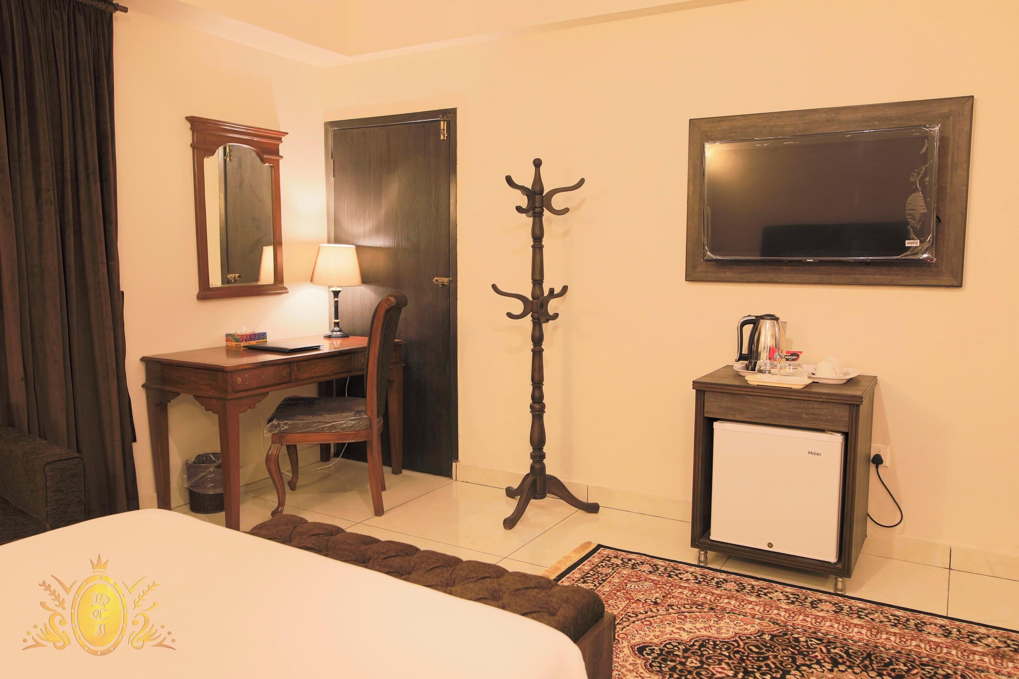 A hotel room with a bed, a desk with a chair, a coat stand, a small refrigerator, and a mirror on the wall.