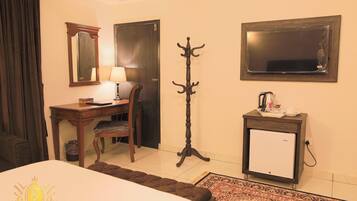 A hotel room with a bed, a desk with a chair, a coat stand, a small refrigerator, and a mirror on the wall.