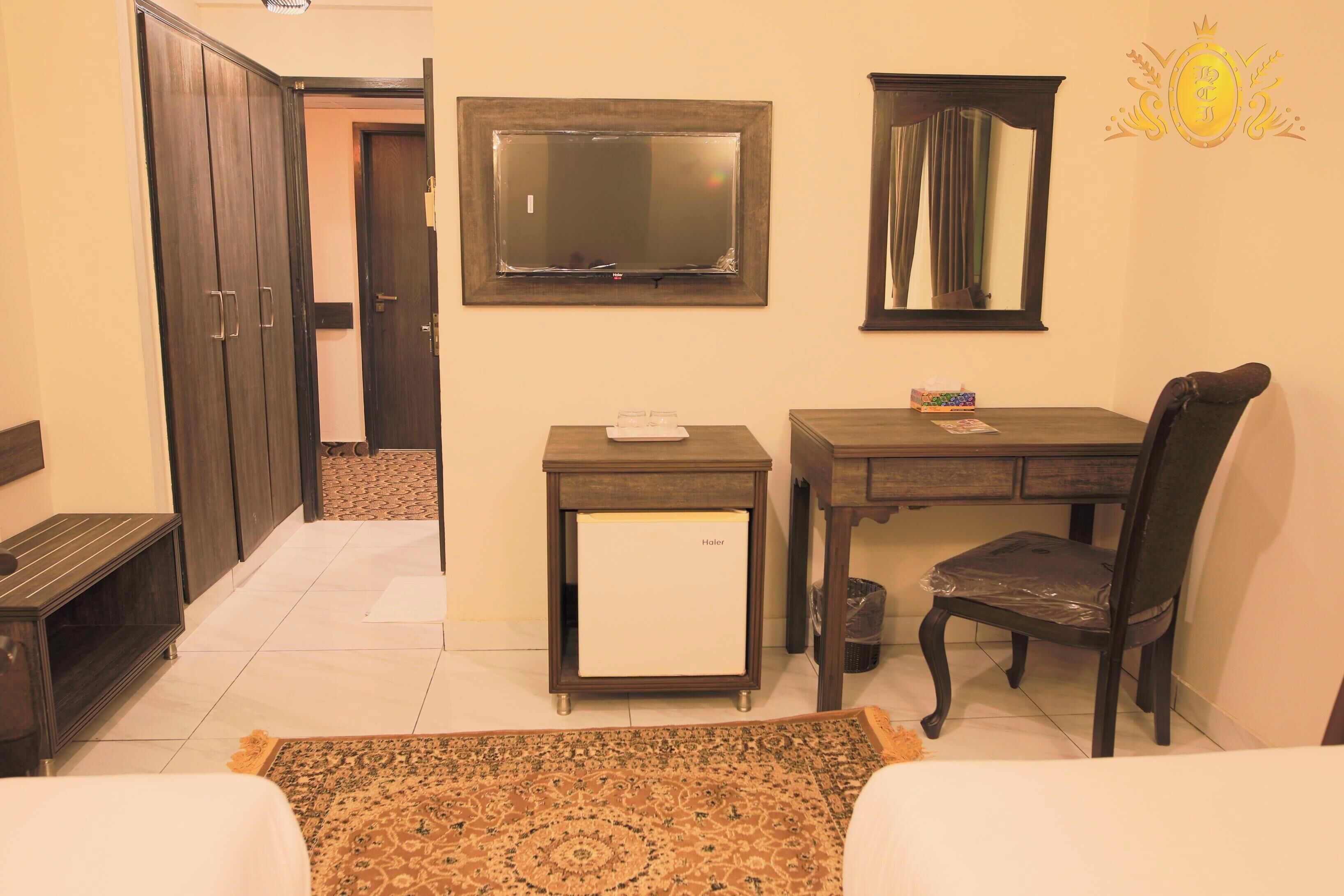 A hotel room with a bed, a desk, a chair, a small refrigerator, a mirror, and a wardrobe.