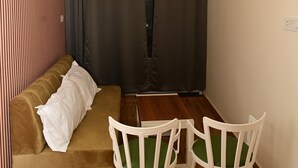 A room with a brown sofa, white cushions, a small table, and two white chairs with green cushions. A ceiling fan and three hanging lights are visible. - Green Park (Varanasi)