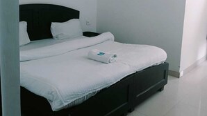 Una cama individual con cabecera oscura, ropa de cama blanca y una toalla doblada con cuidado. Un equipo de aire acondicionado está montado en la pared sobre la cama.