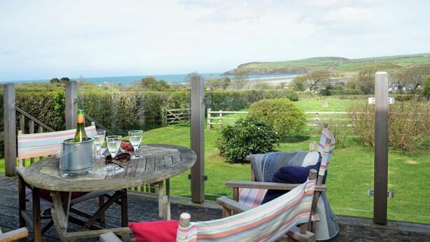 A wooden deck with a table, chairs, and a hammock overlooking a coastal landscape.