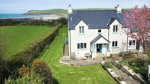 A two-story house with a large lawn, a garden with various plants, and a view of the sea.