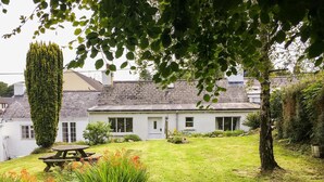 A white house with a sloped roof, a garden with a picnic table, and a tall tree in the foreground.