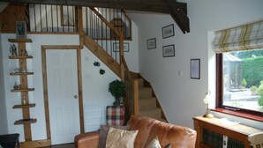 A cozy living room with a brown leather sofa, a wooden staircase, a bookshelf, and a window with a view of greenery.