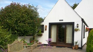 A white building with a black door, a wooden bench, and a purple folding chair in the courtyard.