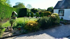 A well-maintained garden with neatly trimmed hedges, a small gazebo, and a paved pathway leading to a white building with a picture on the wall.
