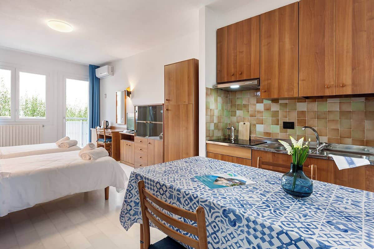 A compact hotel room with two beds, a dining table, a kitchenette, and a view of greenery outside.