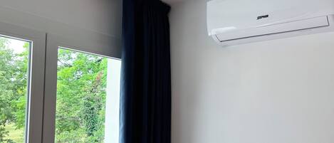 Una habitación con un equipo de aire acondicionado blanco, marcado con 'ZEPHIR', montado en la pared, una ventana que da a vistas de vegetación y una cortina oscura parcialmente bajada.