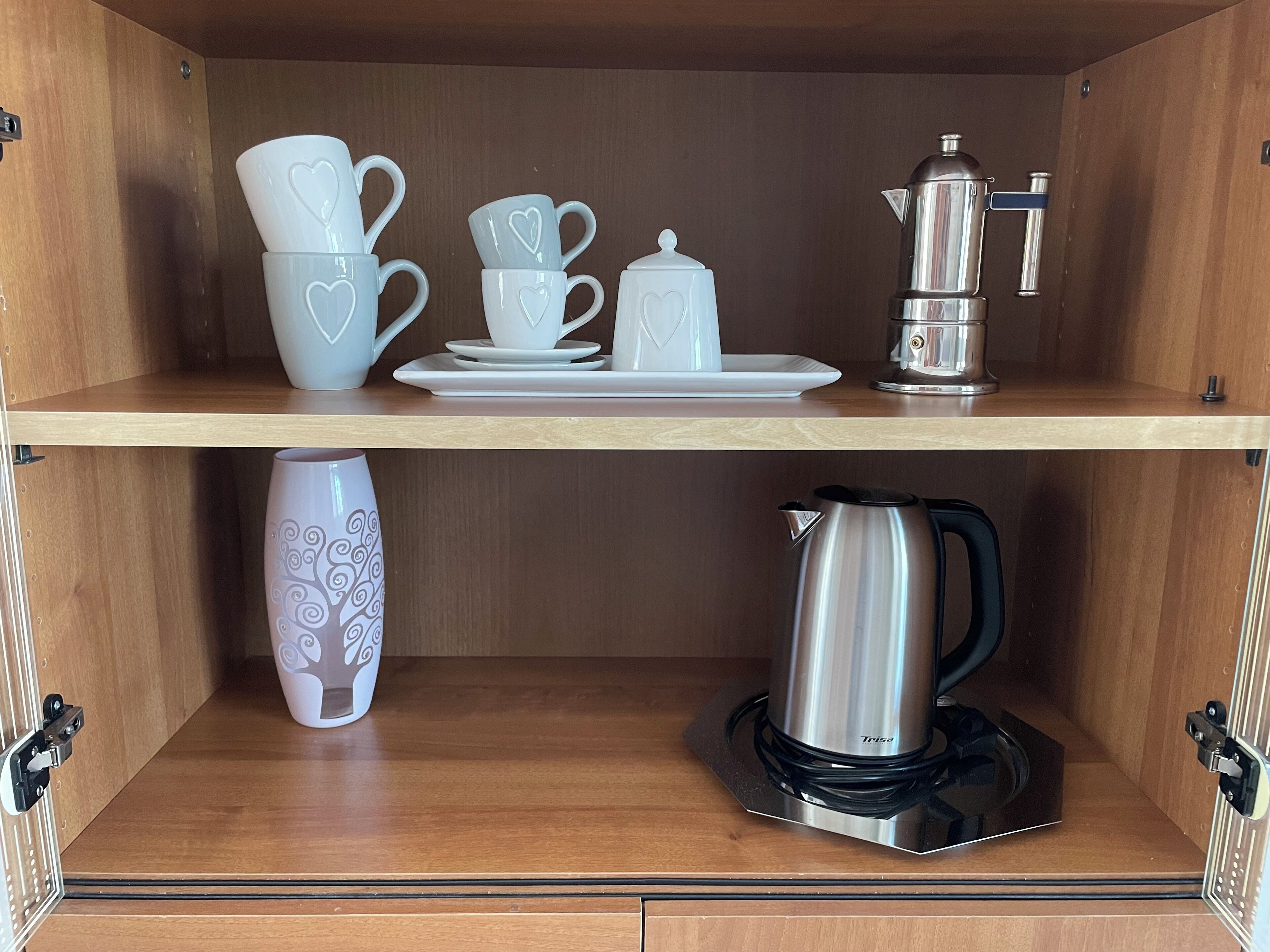 A kitchen with a coffee maker, a kettle, and various white cups and saucers on shelves.