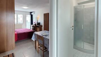 A small room with a bed, a desk, a chair, a table, and a shower.