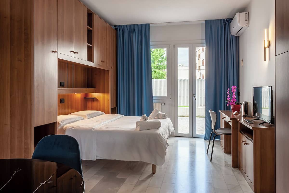 A hotel room with a bed, a desk, a chair, and a TV. There is a window with curtains, and an air conditioning unit on the wall.