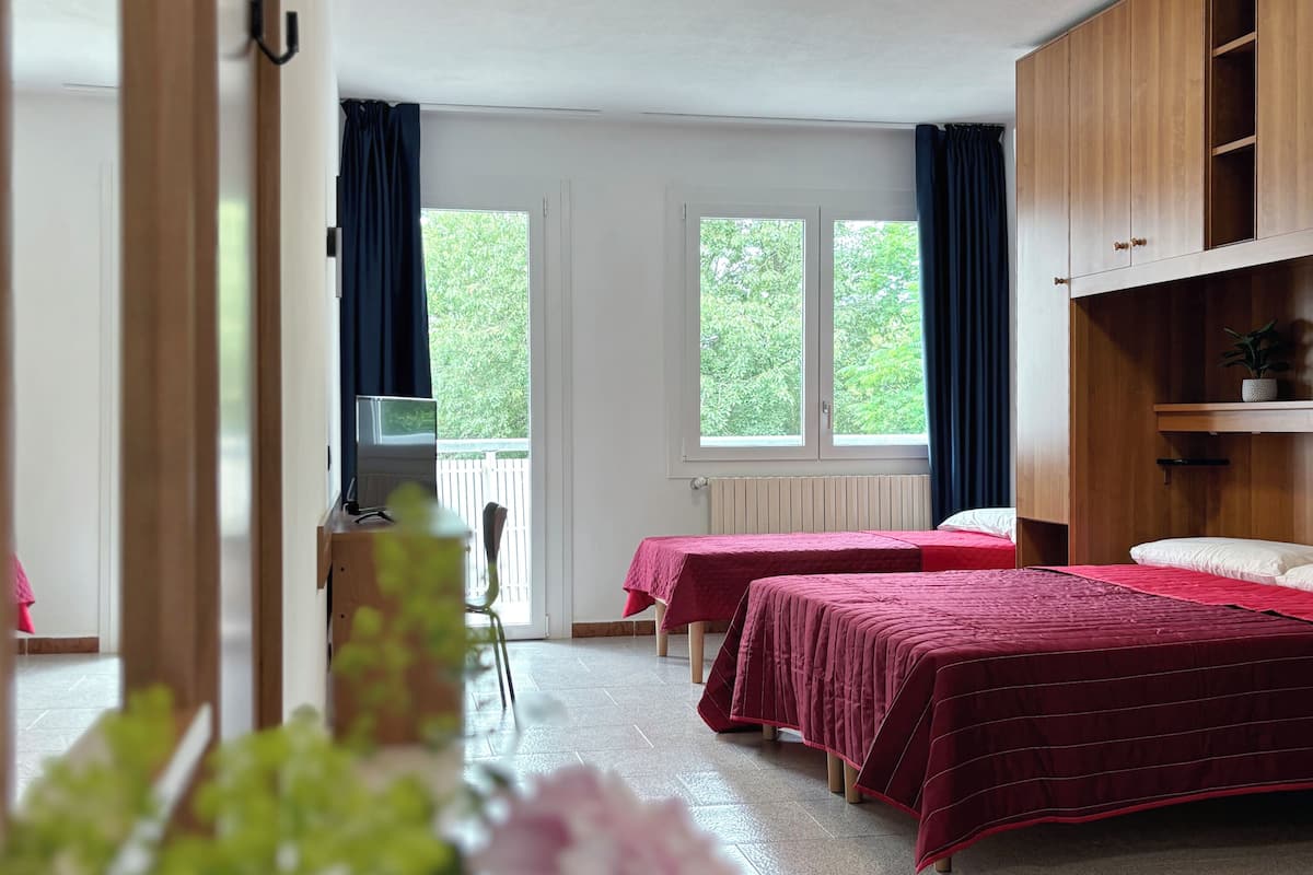 A hotel room with two beds, a wooden wardrobe, a desk, and a window with curtains.
