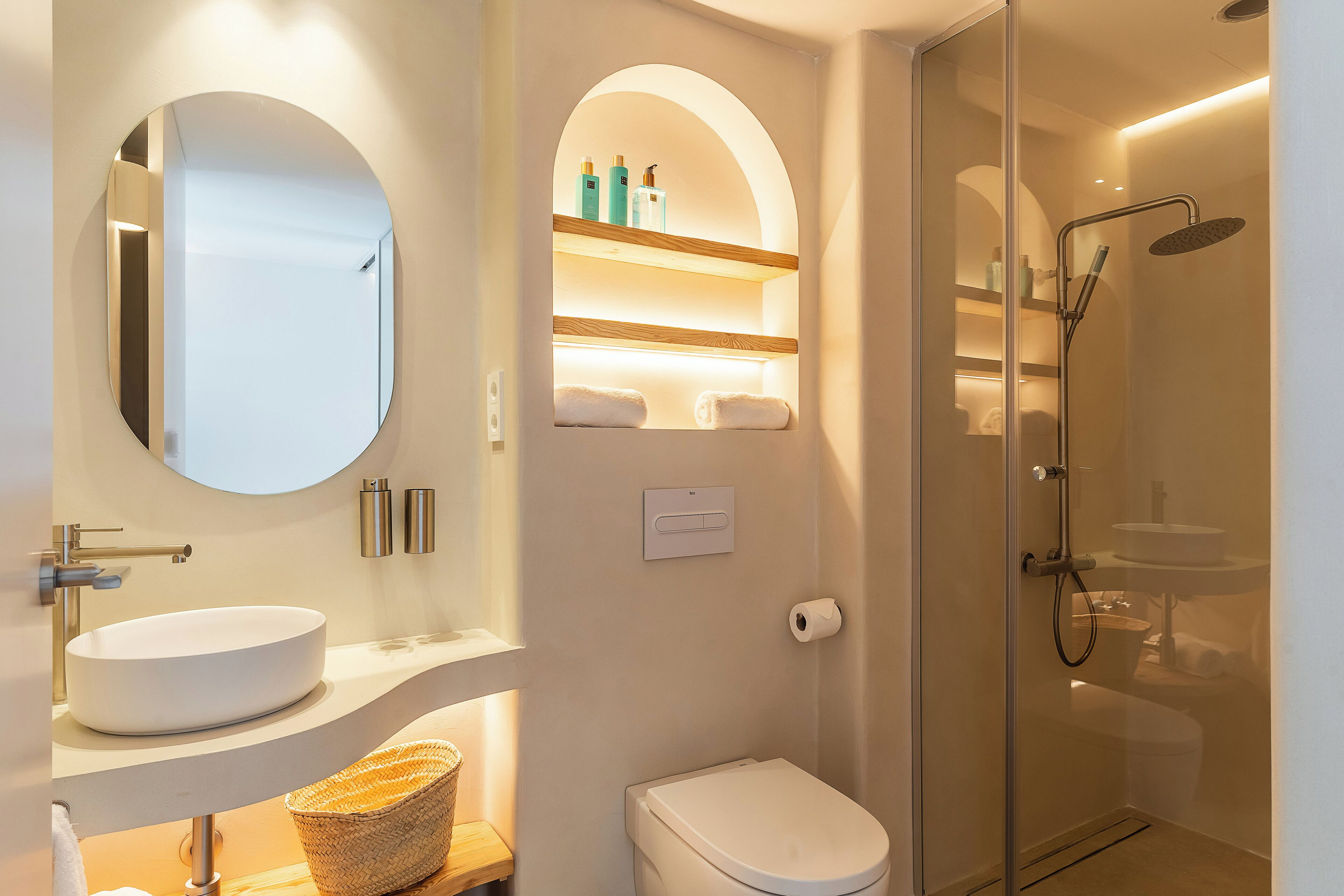 A modern bathroom with a round mirror, a white sink, a toilet, and a glass-enclosed shower area.