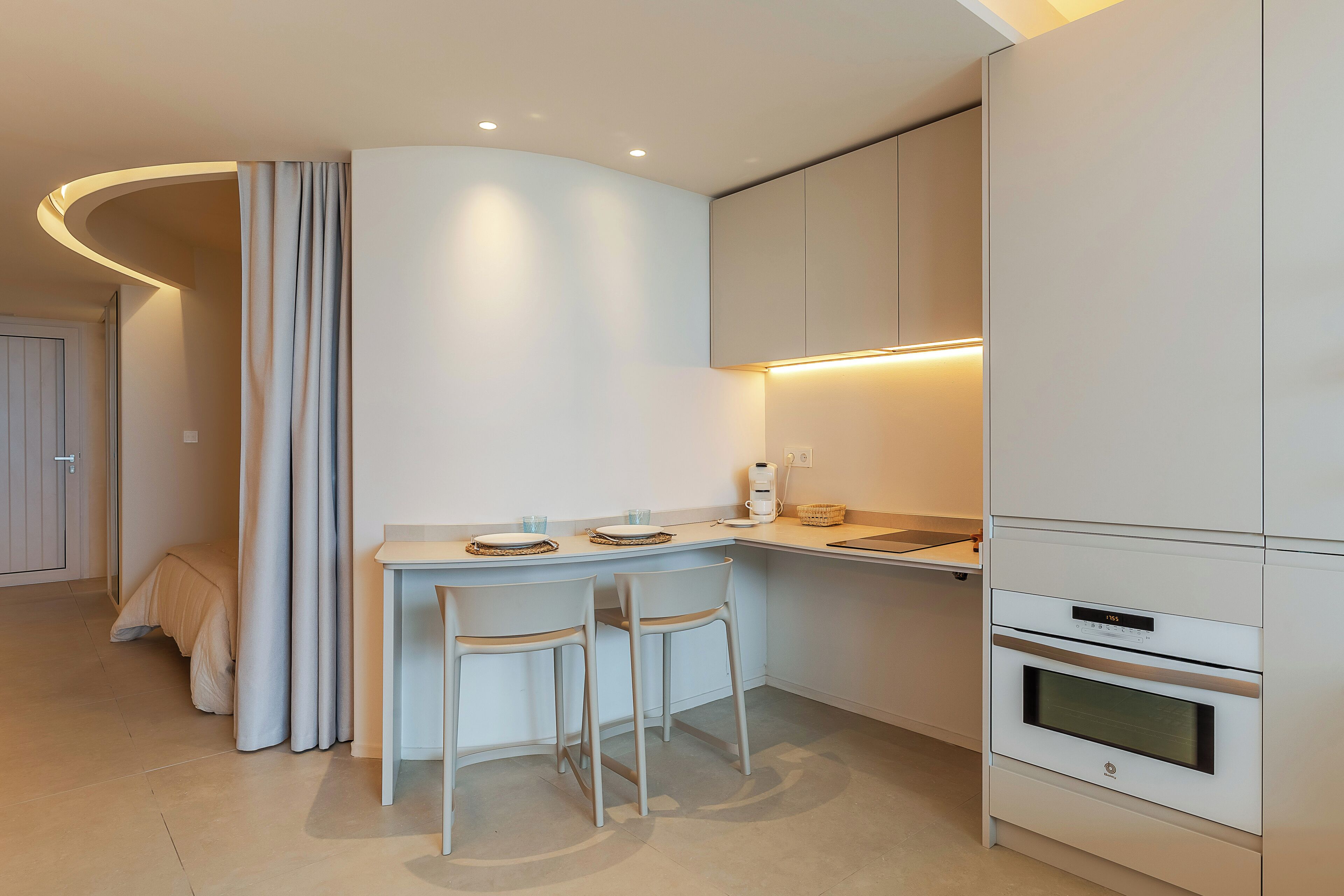 Una cocina moderna con una encimera curva, dos sillas blancas, horno integrado y un pequeño comedor con mesa y sillas.