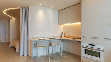 Una cocina moderna con una encimera curva, dos sillas blancas, horno integrado y un pequeño comedor con mesa y sillas.