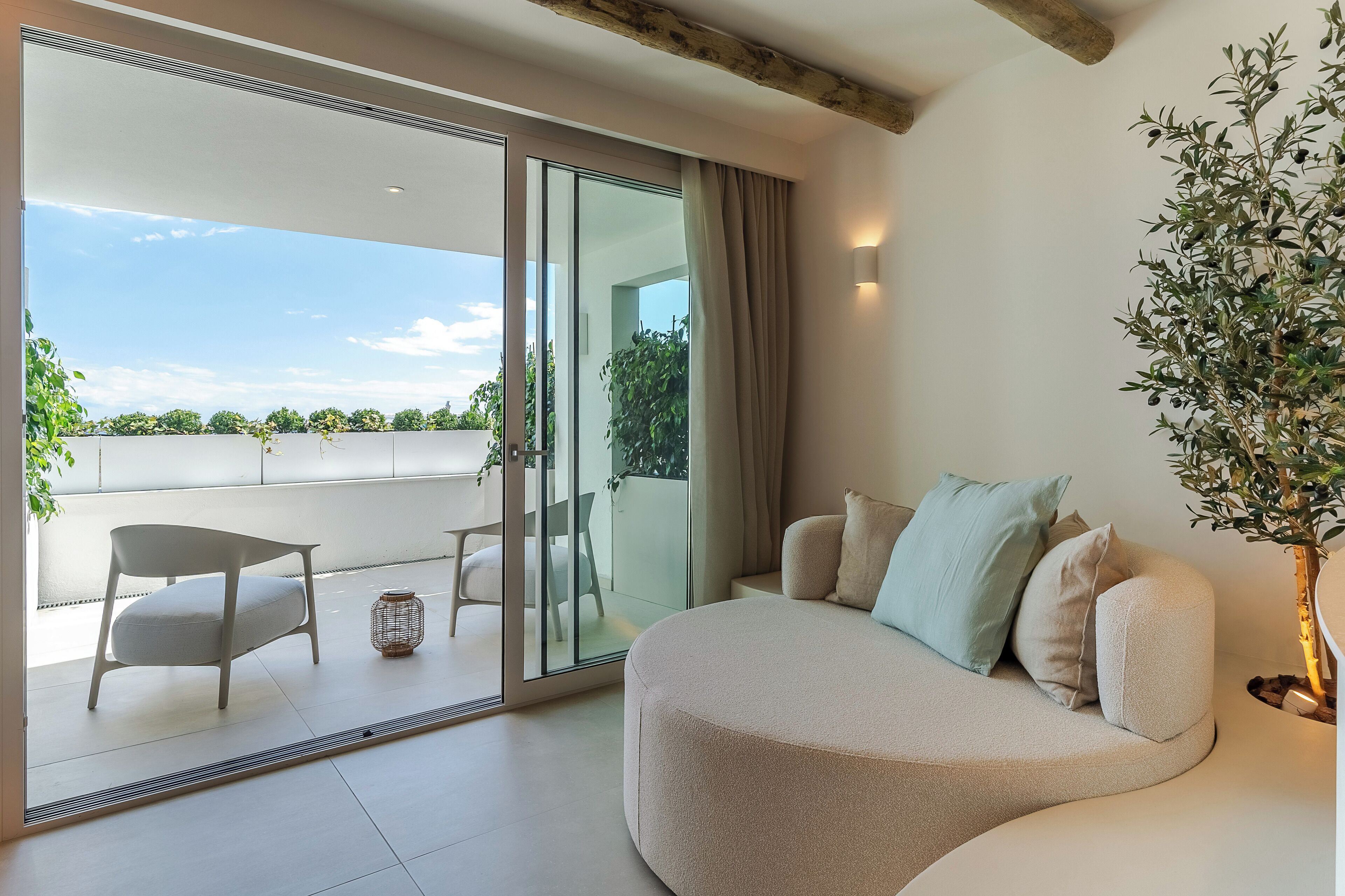 Una sala de estar moderna con un sofá curvo, una mesita y una silla. Hay un ventanal corredero que da a un balcón con vistas al cielo y al follaje.