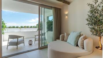 Una sala de estar moderna con un sofá curvo, una mesita y una silla. Hay un ventanal corredero que da a un balcón con vistas al cielo y al follaje.