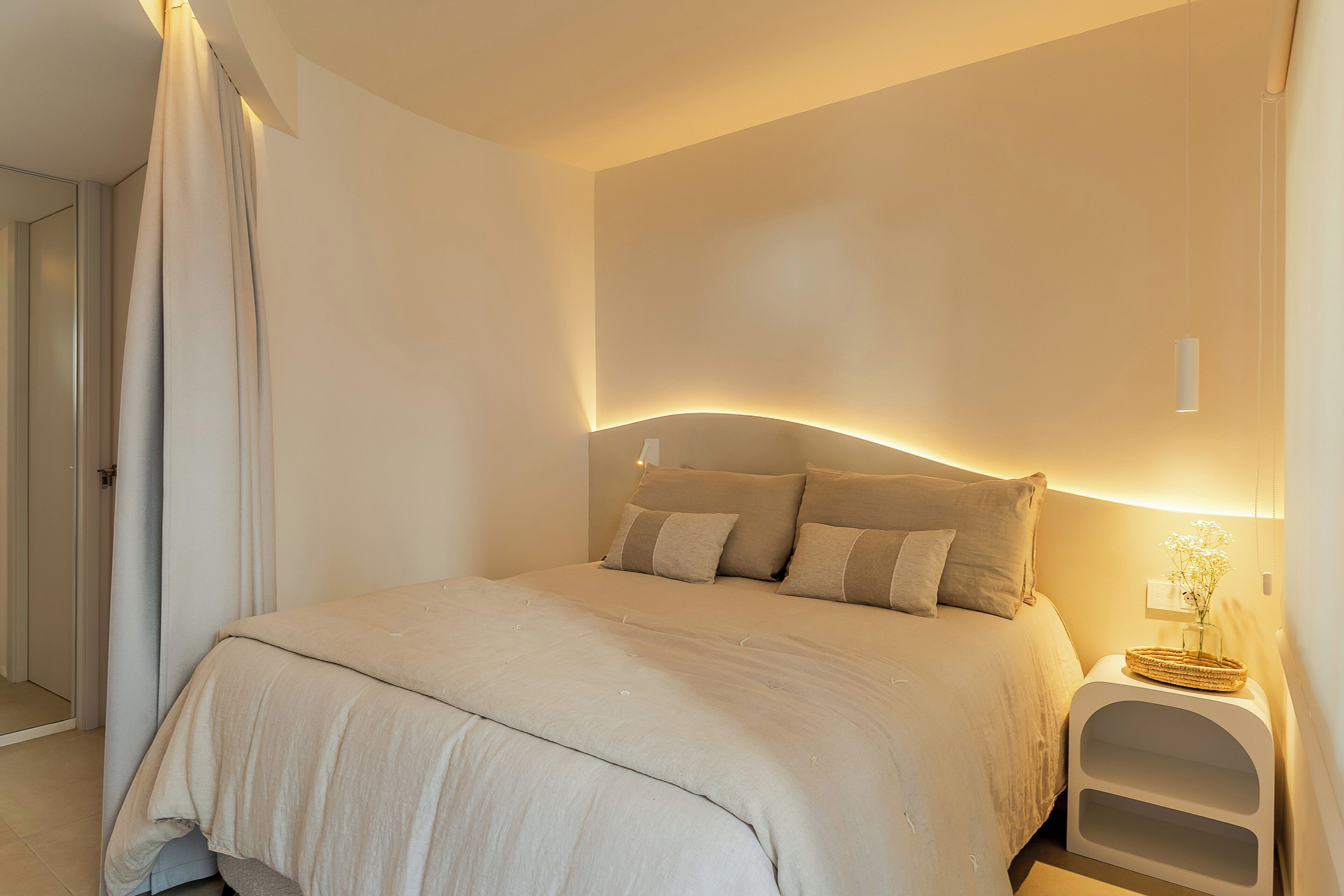 Una cama bien hecha con cabecera, una mesita de noche con un jarrón de flores y una lámpara fijada a la pared.