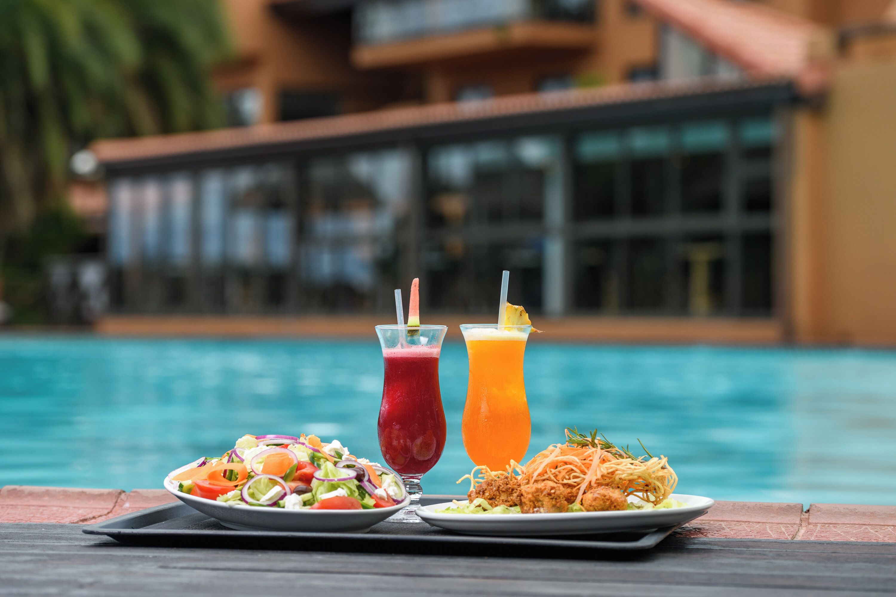 Uma mesa de jantar com uma salada, uma bebida vermelha e uma bebida amarela perto de uma piscina.