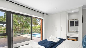 Et moderne hotelværelse med en stor skydedør, der fører til et poolområde, en seng med hvide sengetøj, en blå accentvæg og en lille træbænk.
