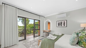 Et moderne soveværelse med en stor seng, udsigt til grøn natur, en skydedør af glas og et lille skrivebord med lampe.