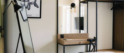 A modern hotel room with a bed, a desk with a chair, a hanging light fixture, and a framed picture on the wall.