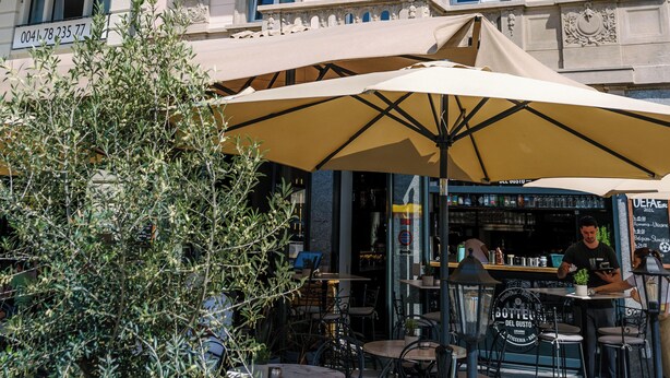 Un edificio de varios pisos con balcones ornamentales y una zona de café con mesas al aire libre.