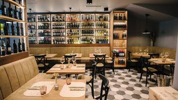 Un restaurante con varias mesas y cabinas, cada una con un mantel individual, cubiertos y una copa. Las paredes están revestidas de estanterías que exhiben diferentes botellas y vasos.