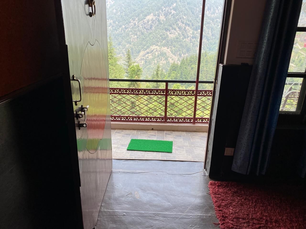 Een geopende deur die leidt naar een balkon met een groene mat, een metalen hekwerk en uitzicht op een bergachtig landschap.