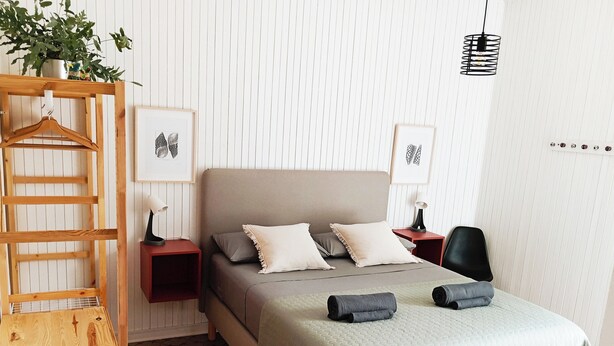 Une chambre à coucher bien rangée, avec un lit, des tables de chevet, une chaise en bois, une plante suspendue et des œuvres d’art accrochées aux murs.