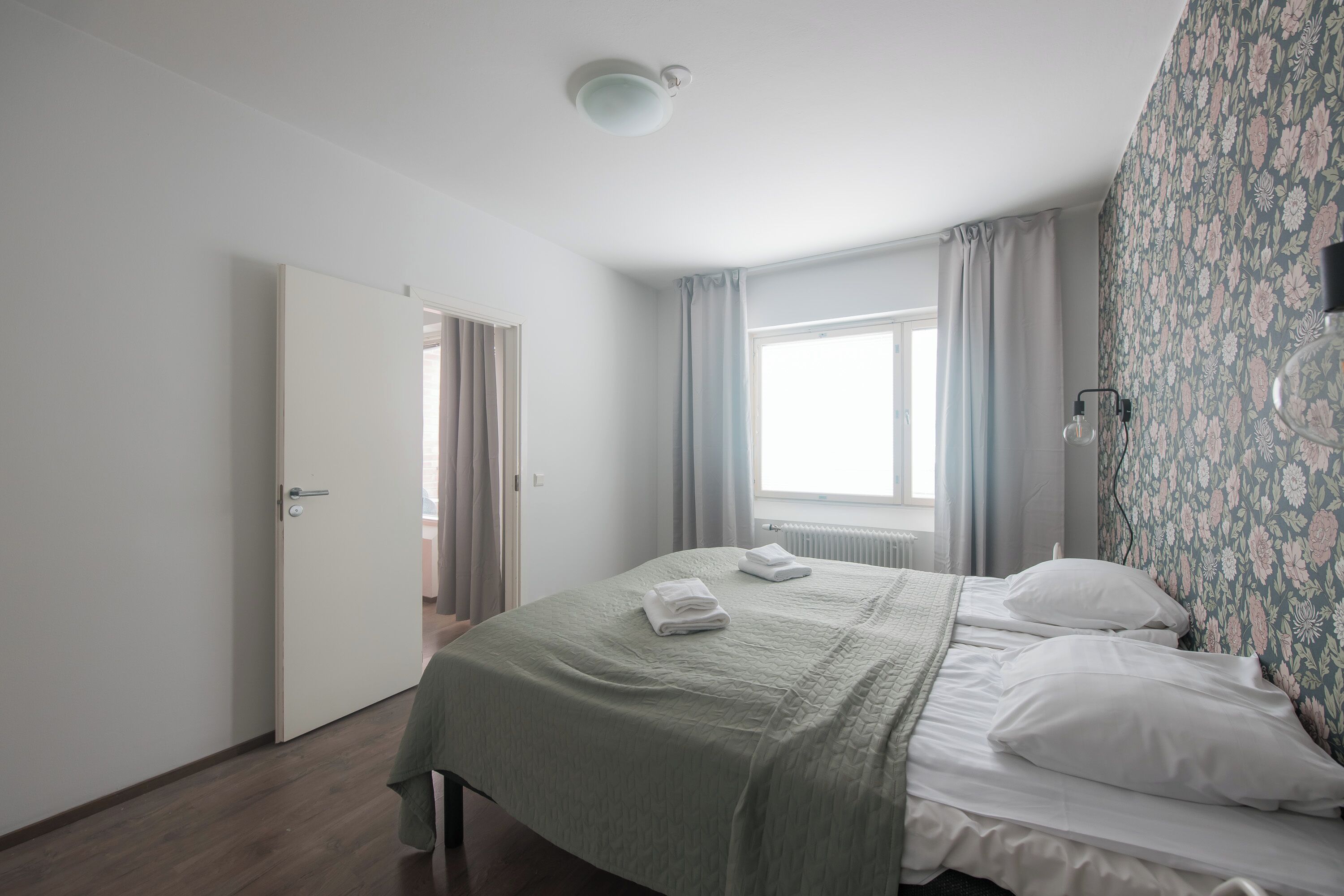 Une chambre à coucher avec un lit, une porte, une fenêtre avec des rideaux et un mur orné d’un motif floral.