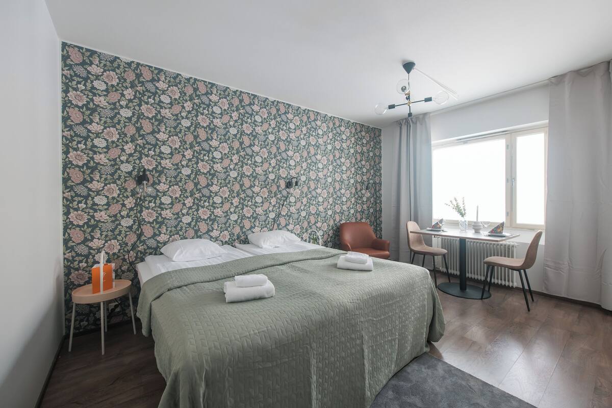 A bedroom with a floral wallpaper, a bed with green bedding, a round table with chairs, and a window with curtains.