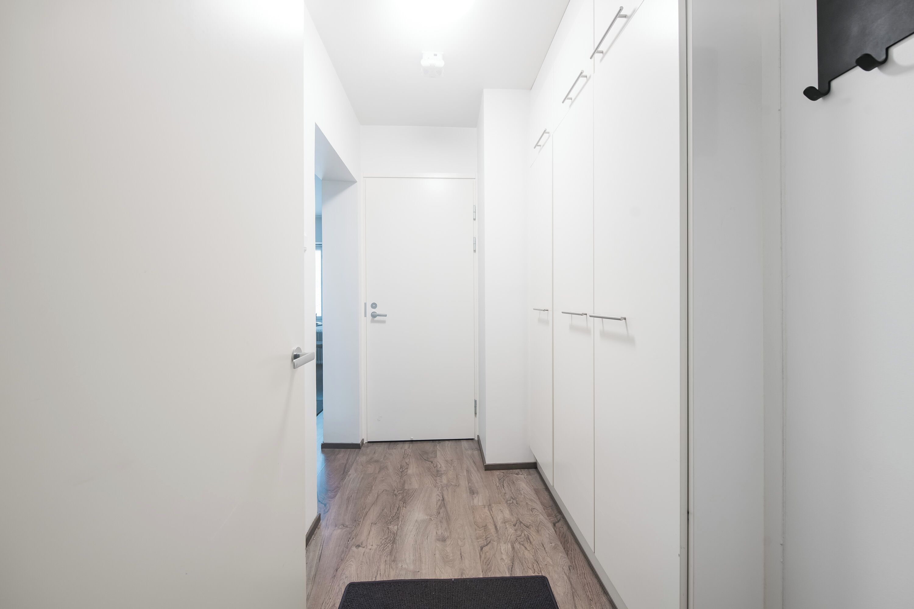 A narrow hallway with white walls, a wooden floor, and a closed door at the end.