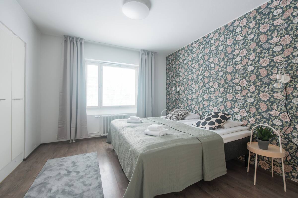 A bedroom with a bed, a window, a radiator, a plant, and a wall with floral wallpaper.