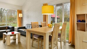 A modern living room with a wooden dining table, chairs, and a yellow pendant light. There is a black leather sofa and a small wooden cabinet with shelves.