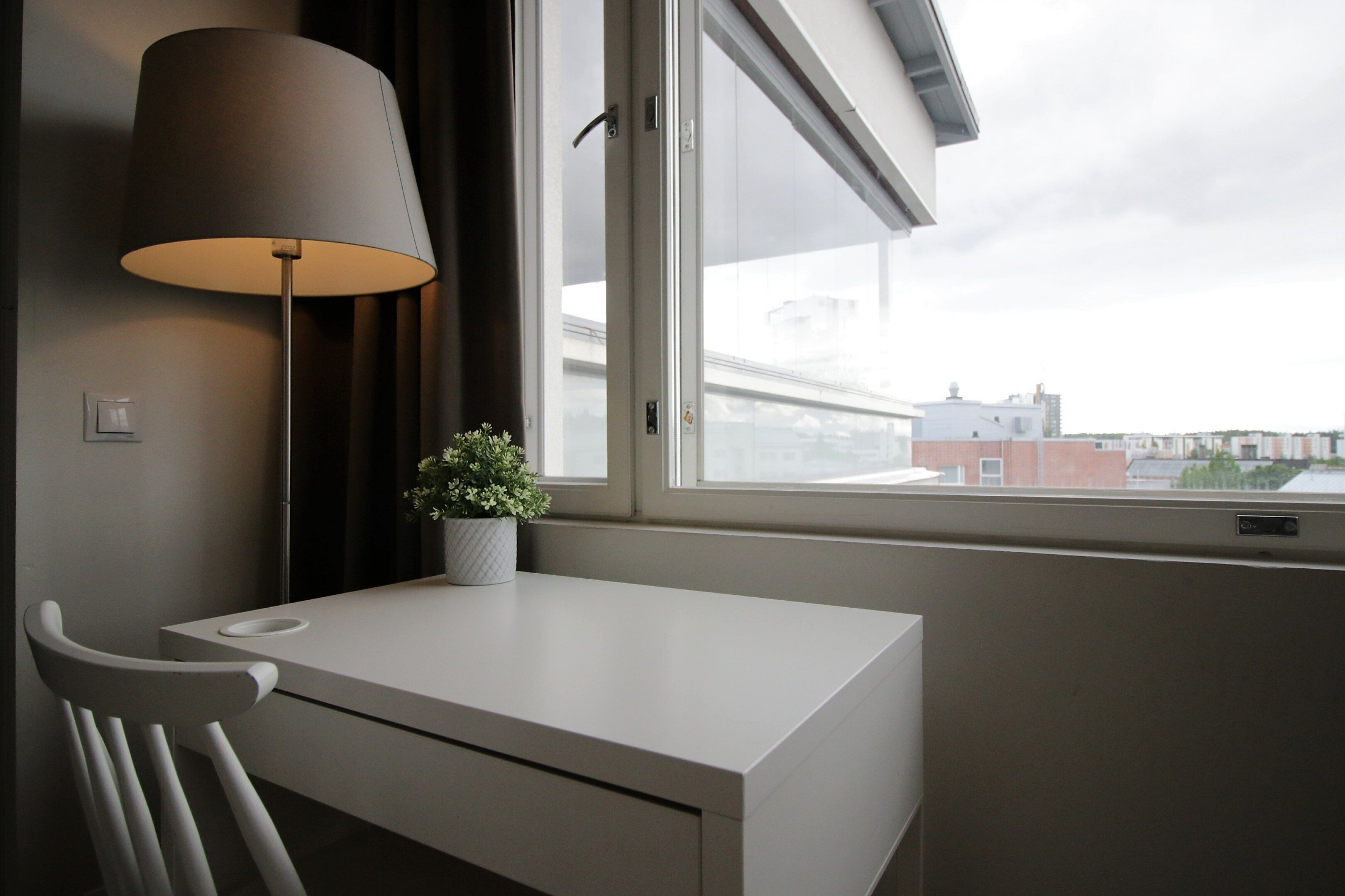 Ett modernt rum med ett vitt skrivbord, en lampa, en krukväxt och en utsikt över byggnader genom ett fönster.