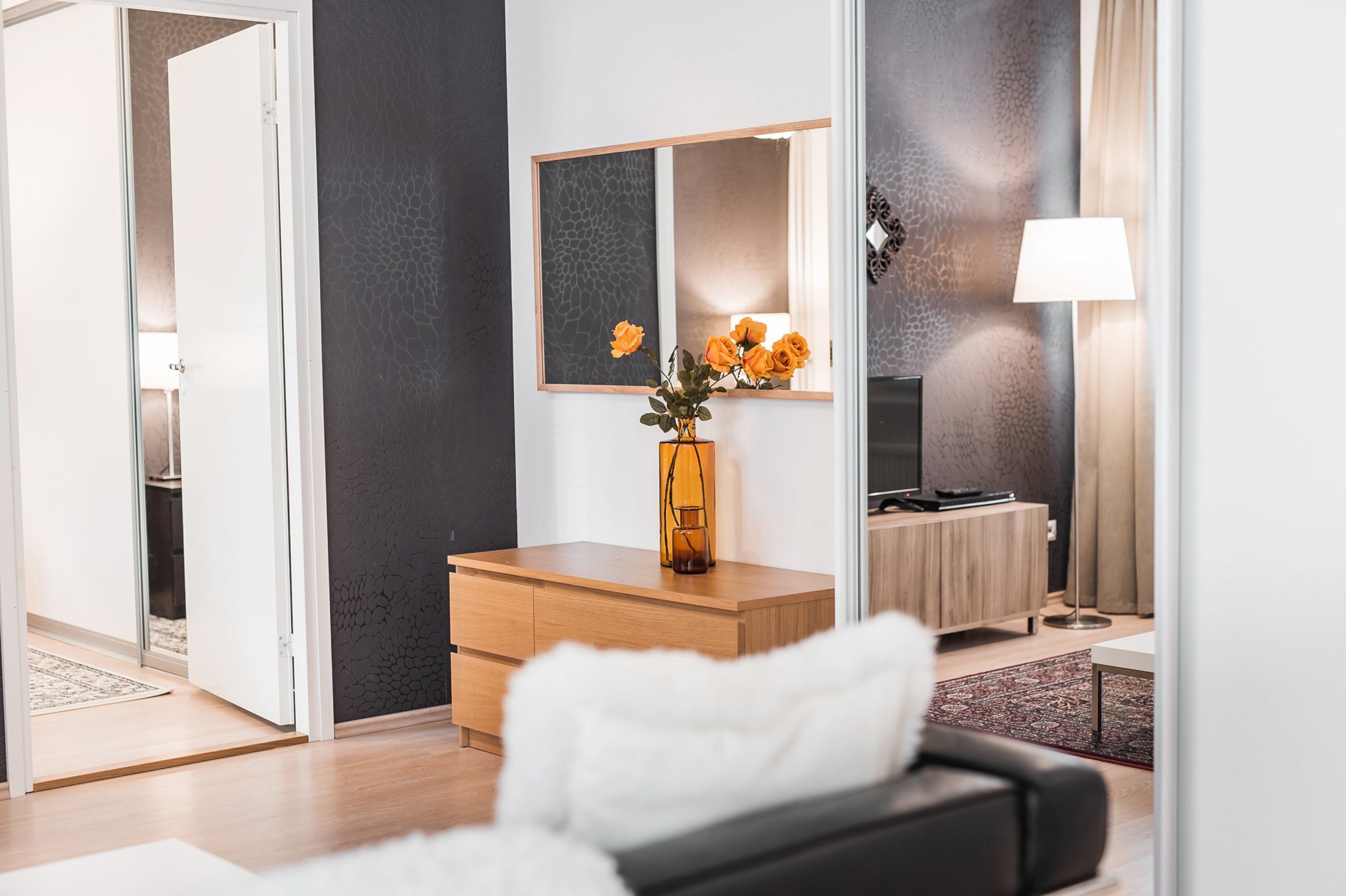 A modern living room with a wooden cabinet, a mirror, a vase with flowers, a TV, and a lamp.