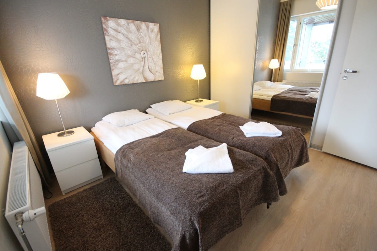 Habitación de hotel con dos camas, una mesita de noche con lámpara, un radiador y un cuadro en la pared.