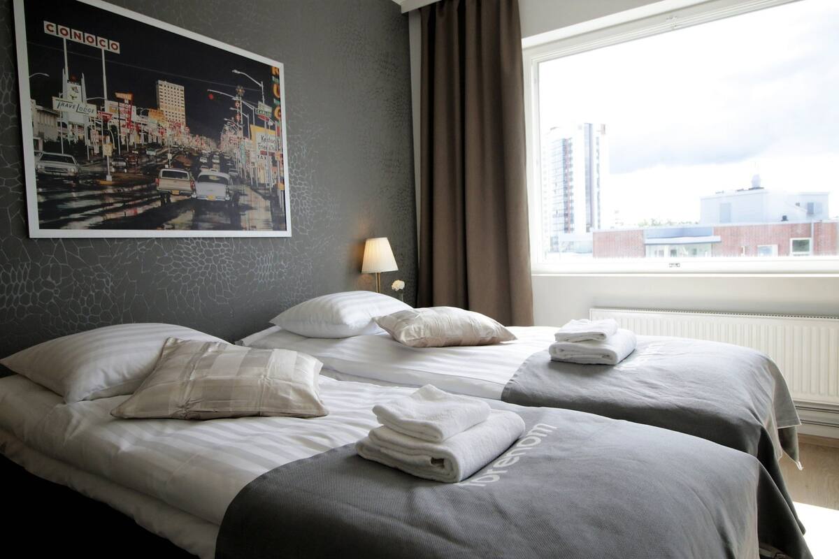 Habitación de hotel con dos camas, un cuadro de paisaje urbano, una ventana con cortinas y una lámpara.
