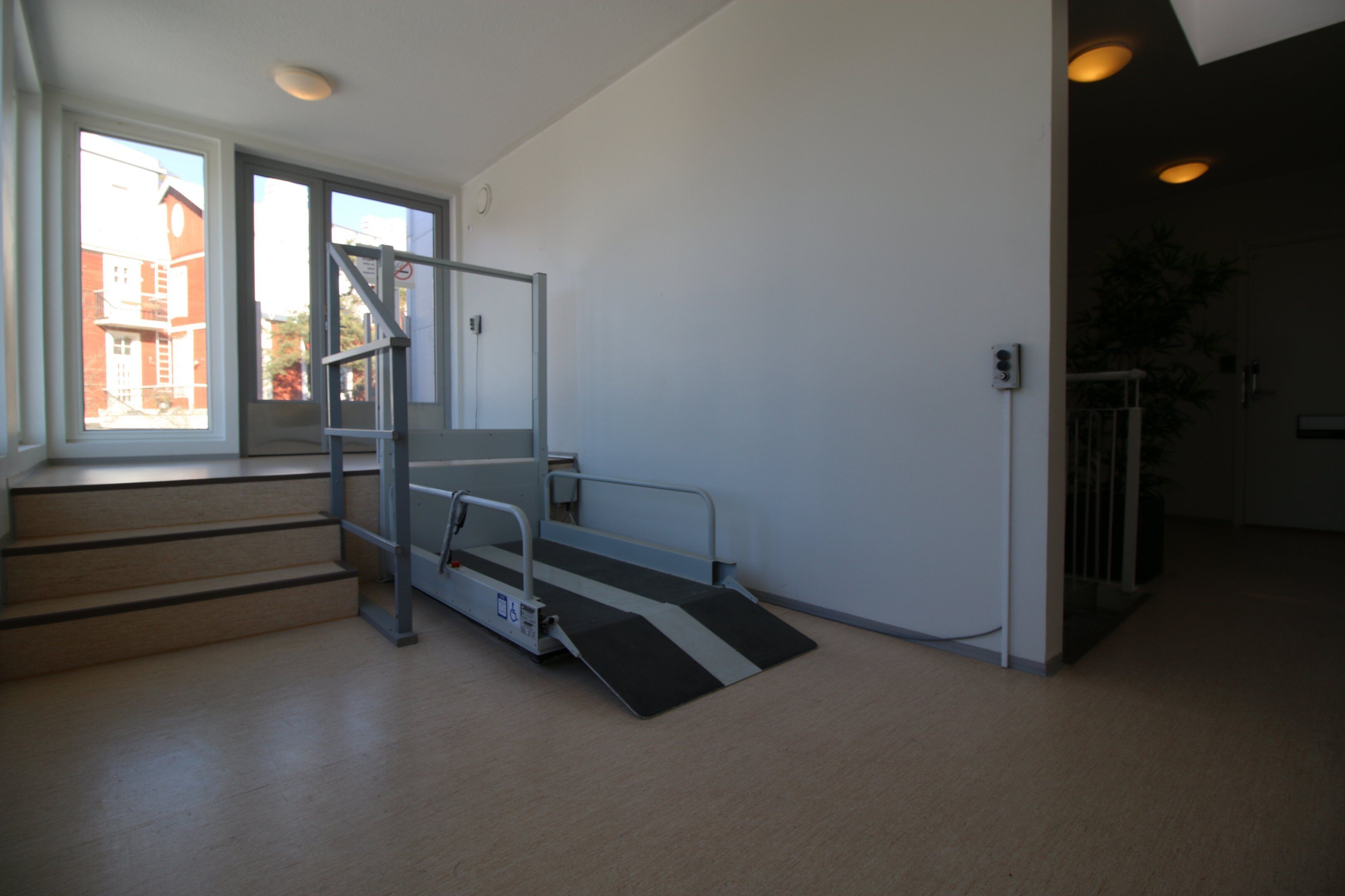 A room with a wheelchair ramp at the entrance, a staircase, and a view of buildings outside.